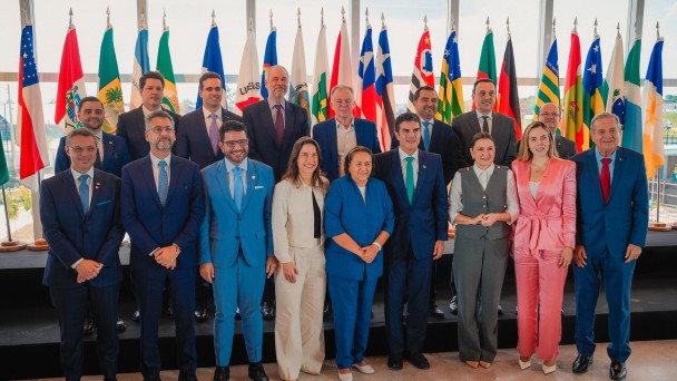 A imagem mostra um grupo de cerca de 20 pessoas, homens e mulheres, de pé em três fileiras. Eles estão vestidos com roupas sociais, como ternos e tailleurs. A maioria dos homens veste ternos em tons de azul ou cinza escuro, enquanto as mulheres usam tailleurs em cores como branco, azul claro e rosa. A pessoa em destaque no centro da primeira fila é um homem alto com cabelo escuro, vestindo um terno azul-celeste. Ele está sorrindo. Atrás do grupo, há uma fileira de mastros com bandeiras de diferentes estados brasileiros e a bandeira do Brasil. O ambiente parece ser um espaço de reunião ou evento oficial, com iluminação clara.