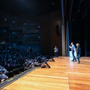 Auditório lotado com várias pessoas assistindo a uma apresentação. Três pessoas estão no palco de madeira, uma fala ao microfone. Plateia atenta, alguns registros com celular. Ambiente amplo e com vários níveis de assentos.