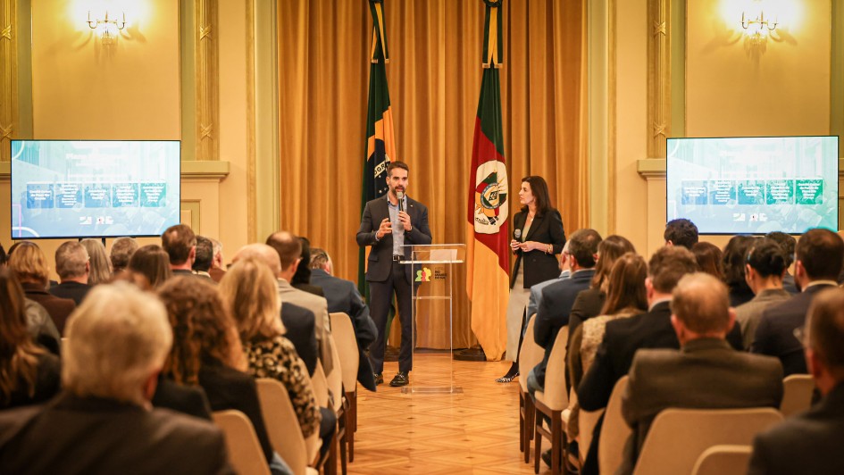 A imagem retrata uma sala onde diversas pessoas estão sentadas, assistindo a uma apresentação. No centro, o governador fala em um púlpito transparente, acompanhado pela titular da Sict, que segura um microfone. Atrás deles, as bandeiras do RS e do Brasil estão expostas. Nas laterais do ambiente, duas telas grandes exibem os slides da apresentação.