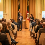 A imagem retrata uma sala onde diversas pessoas estão sentadas, assistindo a uma apresentação. No centro, o governador fala em um púlpito transparente, acompanhado pela titular da Sict, que segura um microfone. Atrás deles, as bandeiras do RS e do Brasil estão expostas. Nas laterais do ambiente, duas telas grandes exibem os slides da apresentação.