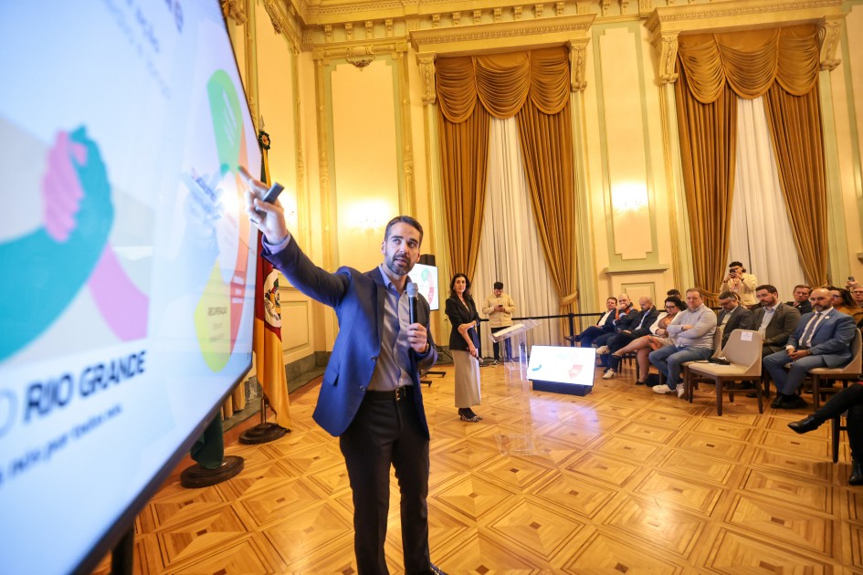 A imagem mostra o governador apresentando diante de uma tela grande com gráficos coloridos. Ele segura um microfone e aponta para a tela, enquanto o público, sentado em cadeiras, acompanha atentamente.