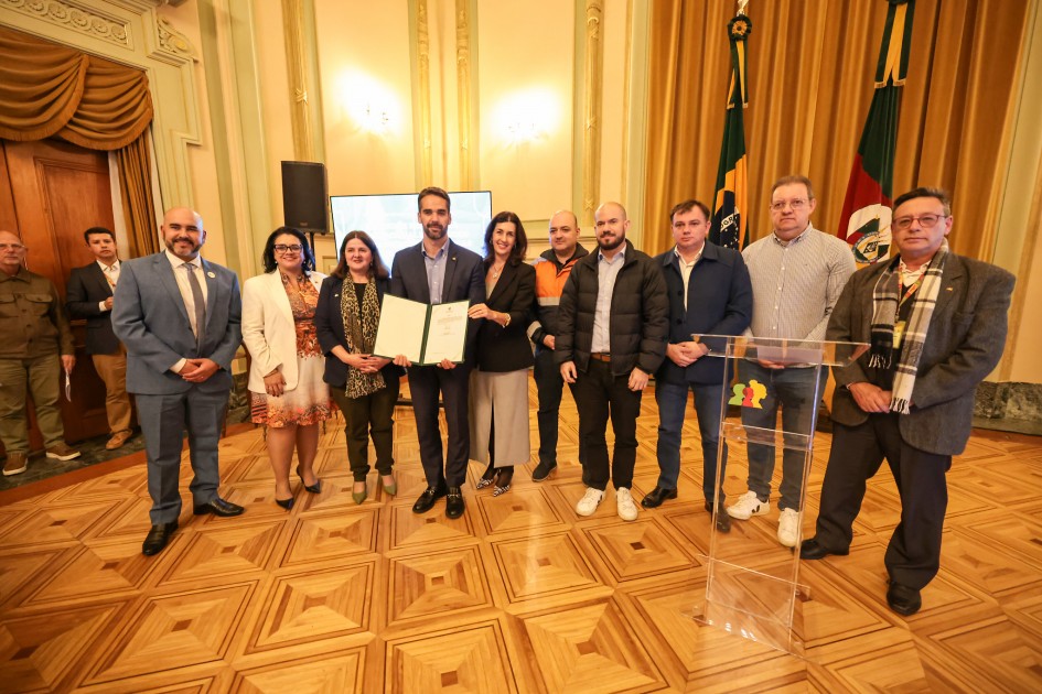A imagem mostra um grupo de pessoas em um ambiente formal, com o governador Eduardo Leite ao centro exibindo um documento aberto. Há um púlpito com microfones à direita, bandeiras ao fundo e decoração elegante.