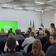 Esta é uma fotografia de um evento formal em um auditório, visto da parte de trás da plateia, olhando em direção ao palco.

No canto superior esquerdo, um homem de terno escuro está em pé ao lado de um telão verde-claro. O telão exibe o texto "Assinatura de convênios". O homem gesticula com as mãos enquanto fala em um microfone que está em um pedestal.

A plateia, composta por dezenas de pessoas, está sentada em cadeiras escuras, voltada para o palco. No lado direito do palco, há duas bandeiras de mastros altos, uma do Brasil e outra do Rio Grande do Sul.

As paredes da sala são de cor clara, e o teto é baixo, com luminárias embutidas. A iluminação é uniforme. O ambiente parece ser um evento de assinatura de documentos ou uma reunião oficial.