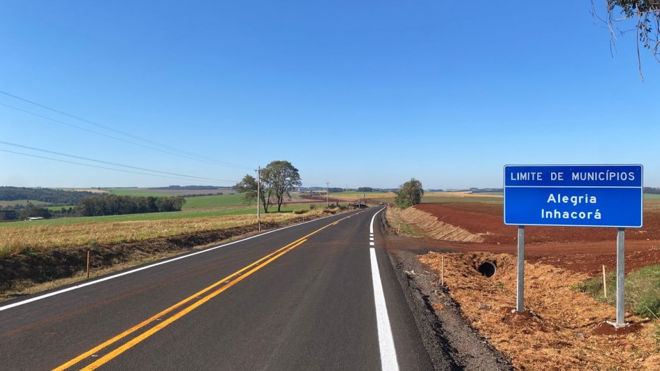 Imagem de uma estrada asfaltada, com faixas centrais amarelas e laterais brancas, que se estende em direção ao horizonte em meio a uma paisagem rural. À direita, há uma placa azul indicando o limite entre os municípios de Alegria e Inhacorá. O céu está limpo e azul, e há campos cultivados dos dois lados da via.