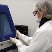 A foto mostra uma mulher de perfil, em um laboratório. Ela usa um jaleco branco, óculos, luvas e uma máscara de proteção. A mulher está em frente a uma máquina de cor azul e branca. A máquina parece ser um analisador genético. A mulher está tocando no painel frontal da máquina, com uma expressão focada.