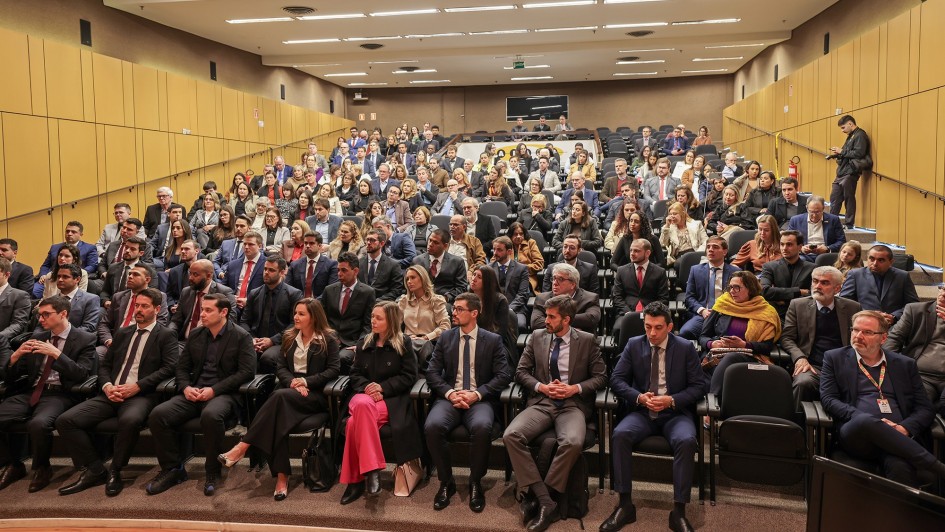 Esta é uma fotografia de um grande auditório em perspectiva, mostrando a plateia sentada e vista do lado do palco.

A plateia, composta por centenas de pessoas, está sentada em cadeiras pretas em filas inclinadas. A maioria dos participantes é adulta e está vestida com trajes formais, como ternos e blazers. Algumas pessoas usam camisas sociais, e há várias mulheres vestindo blazers e camisas sociais.

As paredes laterais do auditório são revestidas com painéis acústicos de cor amarela. O teto é alto e branco, com iluminação embutida. O ambiente é formal e a foto parece ter sido tirada durante um evento, como uma palestra ou uma cerimônia de posse.