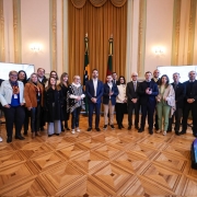 Grupo de pessoas em pé numa sala elegante, com chão de madeira, paredes ornamentadas, bandeiras ao fundo e telas grandes dos dois lados. Iluminação com lustres.