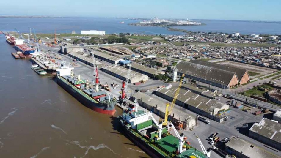 Imagem aérea do Porto do Rio Grande, com grandes navios atracados do lado esquerdo. Ao lado direito, há a estrutura do porto, com sistemas de armazenamento. Ao fundo e ao lado esquerdo, há água. 