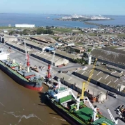 Imagem aérea do Porto do Rio Grande, com grandes navios atracados do lado esquerdo. Ao lado direito, há a estrutura do porto, com sistemas de armazenamento. Ao fundo e ao lado esquerdo, há água. 