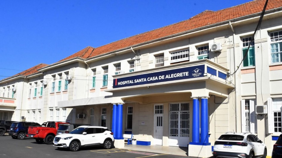 A imagem mostra a fachada principal do Hospital Santa Casa de Alegrete, um prédio de dois andares com paredes claras e telhado de barro avermelhado. A entrada principal, que fica no centro, possui uma marquise sustentada por duas colunas cilíndricas pintadas de azul, e sobre ela há uma placa com os dizeres "HOSPITAL SANTA CASA DE ALEGRETE" e o logotipo do hospital. Veículos brancos e vermelhos estão estacionados em frente. A foto foi tirada em um dia ensolarado, sob céu azul.