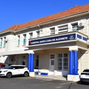 A imagem mostra a fachada principal do Hospital Santa Casa de Alegrete, um prédio de dois andares com paredes claras e telhado de barro avermelhado. A entrada principal, que fica no centro, possui uma marquise sustentada por duas colunas cilíndricas pintadas de azul, e sobre ela há uma placa com os dizeres "HOSPITAL SANTA CASA DE ALEGRETE" e o logotipo do hospital. Veículos brancos e vermelhos estão estacionados em frente. A foto foi tirada em um dia ensolarado, sob céu azul.