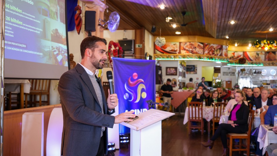 A imagem mostra o governador falando ao microfone em um púlpito, diante de uma plateia sentada em mesas. Ao fundo, há uma tela grande com conteúdo visual e um banner com o logo "ACINP". 