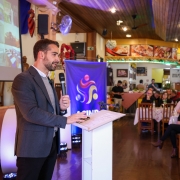 A imagem mostra o governador falando ao microfone em um púlpito, diante de uma plateia sentada em mesas. Ao fundo, há uma tela grande com conteúdo visual e um banner com o logo "ACINP". 