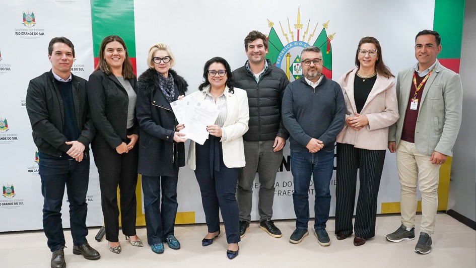 Esta é uma fotografia de um grupo de oito pessoas, quatro homens e quatro mulheres, em pé lado a lado, posando para a câmera.
No centro do grupo, uma mulher de cabelo escuro, com um blazer branco e calça azul-marinho, segura um documento com as duas mãos e sorri. Ao seu lado, à esquerda, uma mulher de cabelo loiro, usando um casaco de lã preto, também sorri. À direita da mulher central, um homem de barba com um suéter e calça jeans está posicionado.
O grupo está diante de uma parede com banners: à esquerda, há um banner vertical com o logotipo e o nome "GOVERNO DO RIO GRANDE DO SUL" em letras pretas sobre fundo verde e branco. À direita, um banner maior, branco e verde, tem o brasão do Rio Grande do Sul e a inscrição "GOVERNO DO ESTADO DO RIO GRANDE DO SUL".
Todos os participantes estão sorrindo. O ambiente é formal e bem iluminado.