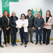 Esta é uma fotografia de um grupo de oito pessoas, quatro homens e quatro mulheres, em pé lado a lado, posando para a câmera.

No centro do grupo, uma mulher de cabelo escuro, com um blazer branco e calça azul-marinho, segura um documento com as duas mãos e sorri. Ao seu lado, à esquerda, uma mulher de cabelo loiro, usando um casaco de lã preto, também sorri. À direita da mulher central, um homem de barba com um suéter e calça jeans está posicionado.

O grupo está diante de uma parede com banners: à esquerda, há um banner vertical com o logotipo e o nome "GOVERNO DO RIO GRANDE DO SUL" em letras pretas sobre fundo verde e branco. À direita, um banner maior, branco e verde, tem o brasão do Rio Grande do Sul e a inscrição "GOVERNO DO ESTADO DO RIO GRANDE DO SUL".

Todos os participantes estão sorrindo. O ambiente é formal e bem iluminado.