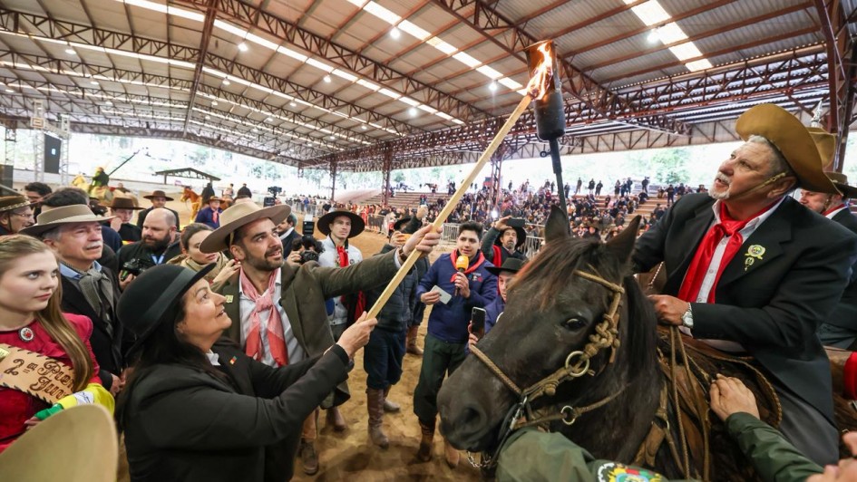 Pessoa a cavalo segura uma tocha enquanto outra encosta um bastão nela, em arena coberta. Público observa; parece cerimônia tradicional.