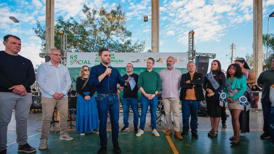 Evento comunitário com várias pessoas; destaque para um orador com microfone em frente a banner “RS Social”.