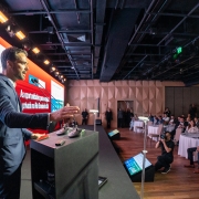 Homem de terno azul escuro fala ao microfone em um palco durante um evento formal, gesticulando com uma das mãos. Ao fundo, há um grande telão colorido com gráficos e textos sobre oportunidades de investimento no Rio Grande do Sul. À direita da imagem, diversas pessoas, vestidas socialmente, estão sentadas em mesas redondas assistindo à apresentação. O ambiente é fechado, com iluminação de auditório e estrutura moderna.