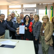 A imagem mostra um grupo de 11 pessoas, em pé, em uma sala de escritório. No centro, uma mulher de blusa preta e um xale vermelho segura um documento em suas mãos, cercada por outras pessoas que também tocam o papel. Os demais indivíduos estão em pé, sorrindo, ao redor. O ambiente tem paredes de madeira e uma grande janela ao fundo. A sala parece ser um escritório do governo, com uma bandeira do Rio Grande do Sul e uma bandeira do Brasil visíveis no canto superior direito.