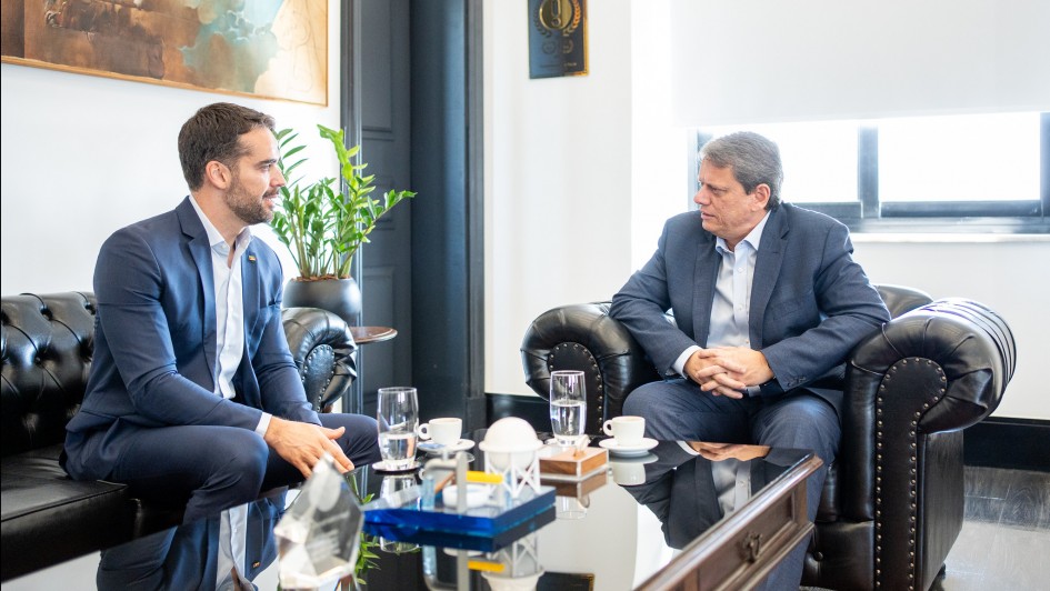A foto mostra o governador Eduardo Leite e o governador Tarcísio de Freitas sentados em poltronas pretas de couro, um de frente para o outro, em uma sala de reunião. Eles estão em uma conversa formal. Eduardo Leite, à esquerda, veste um terno azul-marinho e uma camisa branca, e está olhando para Tarcísio de Freitas. Tarcísio, à direita, usa um terno azul-escuro e uma camisa azul-clara, com as mãos entrelaçadas. Uma mesa de centro escura e baixa separa os dois, com copos de água e xícaras de café sobre ela. O ambiente tem paredes claras e um quadro decorativo de fundo.