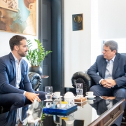 A foto mostra o governador Eduardo Leite e o governador Tarcísio de Freitas sentados em poltronas pretas de couro, um de frente para o outro, em uma sala de reunião. Eles estão em uma conversa formal. Eduardo Leite, à esquerda, veste um terno azul-marinho e uma camisa branca, e está olhando para Tarcísio de Freitas. Tarcísio, à direita, usa um terno azul-escuro e uma camisa azul-clara, com as mãos entrelaçadas. Uma mesa de centro escura e baixa separa os dois, com copos de água e xícaras de café sobre ela. O ambiente tem paredes claras e um quadro decorativo de fundo.