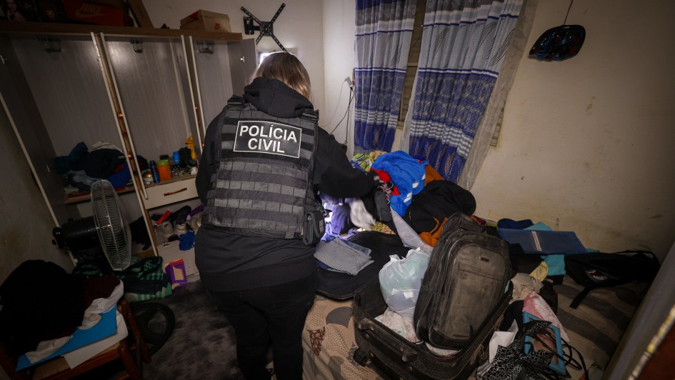 A imagem mostra um quarto escuro e bagunçado, onde uma mulher, de costas para a câmera, está agachada revistando uma cama cheia de roupas e uma mala aberta. Ela usa um colete tático preto com a inscrição "POLÍCIA CIVIL" em letras brancas nas costas. Há um armário e um ventilador à esquerda da imagem. A luz da foto ilumina principalmente a policial, o que realça o ambiente desorganizado. As cortinas da janela são listradas em azul e branco.