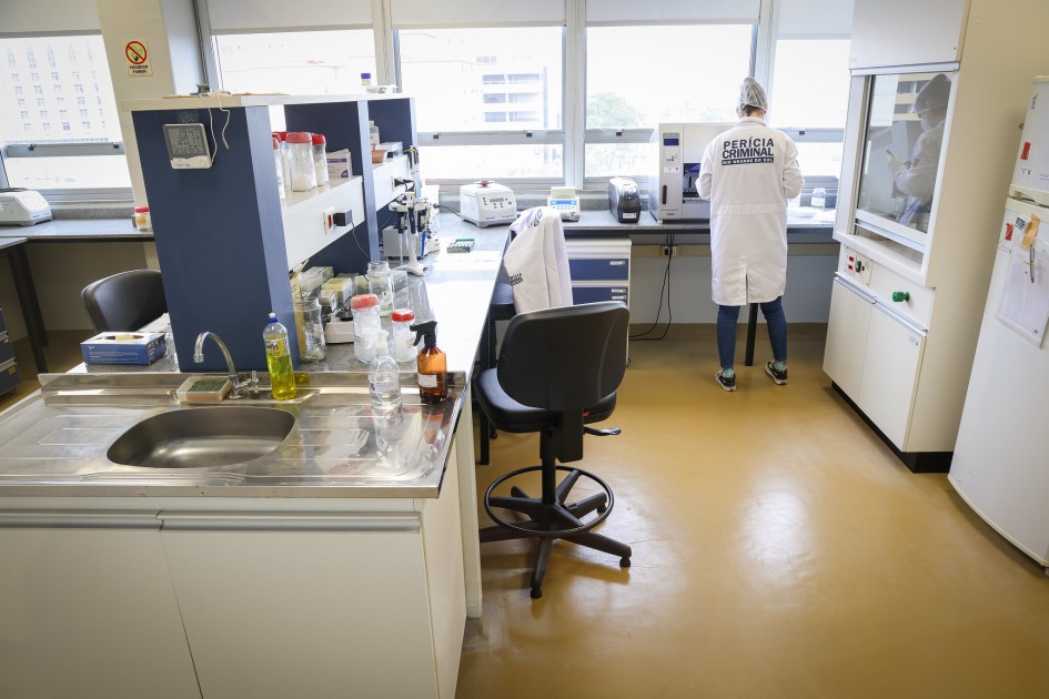 A imagem mostra um laboratório de criminalística bem iluminado. Há uma bancada com uma pia de aço inoxidável em primeiro plano. Ao longo da parede com janelas amplas, há várias estações de trabalho com equipamentos de laboratório. Ao fundo, uma pessoa, vestindo um jaleco branco com a inscrição "PERÍCIA CRIMINAL", está de costas para a câmera, trabalhando em uma cabine de segurança biológica ou um equipamento similar. O chão é de cor clara, e o ambiente é organizado e limpo.