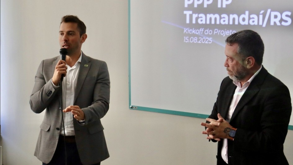 A fotografia mostra dois homens em pé em uma sala de apresentação. O homem à esquerda, com cabelo e barba castanhos, segura um microfone com a mão direita e gesticula com a esquerda enquanto fala. Ele veste um blazer cinza sobre uma camisa branca e calças escuras. O homem à direita, com barba grisalha, está com as mãos unidas à frente do corpo e olha para o chão. Ele usa um paletó escuro e uma camisa rosa.