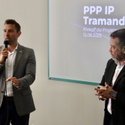 A fotografia mostra dois homens em pé em uma sala de apresentação. O homem à esquerda, com cabelo e barba castanhos, segura um microfone com a mão direita e gesticula com a esquerda enquanto fala. Ele veste um blazer cinza sobre uma camisa branca e calças escuras. O homem à direita, com barba grisalha, está com as mãos unidas à frente do corpo e olha para o chão. Ele usa um paletó escuro e uma camisa rosa.