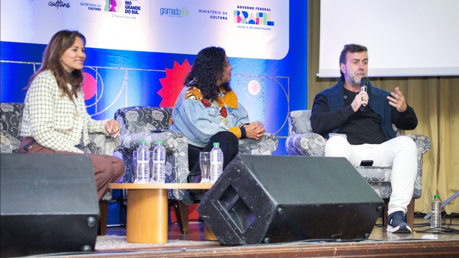 A fotografia mostra três pessoas sentadas em poltronas em um palco, participando de uma discussão em painel. Da esquerda para a direita, estão a presidente da Gramadotur, Rosa Helena Volk, que sorri e veste um blazer xadrez claro. Ao lado dela, a coordenadora institucional do Festival de Cinema de Cartagena, Gisela Perez Fonseca, usa uma jaqueta colorida e olha na direção do orador. Na direita, o diretor-presidente da Embratur, Marcelo Freixo, segura um microfone e gesticula enquanto fala. Ele está de blusa preta e calça branca.