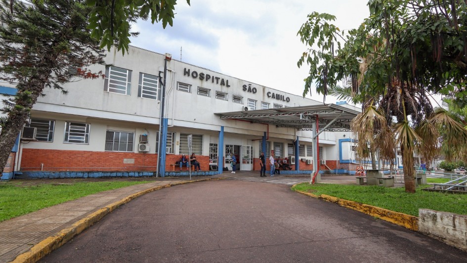 Fachada do Hospital São Camilo de Esteio, com um prédio de dois andares em tons de branco e tijolos aparentes. Na parte superior da edificação, vê-se o letreiro “Hospital São Camilo”. A entrada principal possui uma cobertura metálica, onde algumas pessoas estão sentadas e outras circulam. Há árvores e vegetação ao redor, com caminho asfaltado e calçadas que conduzem até a porta do hospital.