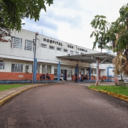 Fachada do Hospital São Camilo de Esteio, com um prédio de dois andares em tons de branco e tijolos aparentes. Na parte superior da edificação, vê-se o letreiro “Hospital São Camilo”. A entrada principal possui uma cobertura metálica, onde algumas pessoas estão sentadas e outras circulam. Há árvores e vegetação ao redor, com caminho asfaltado e calçadas que conduzem até a porta do hospital.
