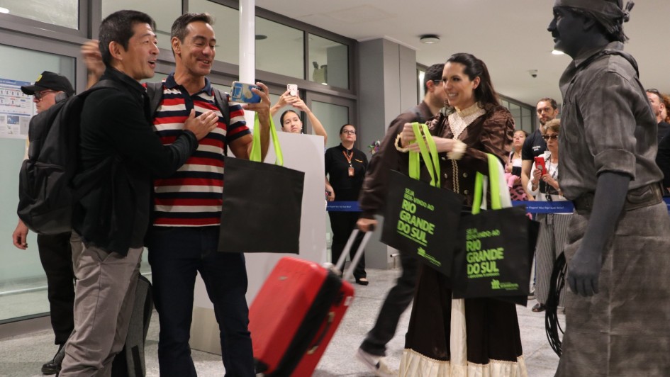 A imagem mostra uma cena em um aeroporto, possivelmente no saguão de desembarque. No centro, um grupo de pessoas está interagindo de forma alegre.

À esquerda, dois homens conversam, sorrindo. Um deles, com traços asiáticos e vestindo uma camisa preta, aponta para algo fora do quadro. O outro, com uma camisa polo listrada em vermelho, branco e azul marinho, está com uma das mãos sobre o peito, em um gesto de surpresa ou gratidão. Ambos seguram sacolas pretas com a frase "RIO GRANDE DO SUL" impressa em letras brancas.

No centro da imagem, uma mulher com um vestido de época marrom e creme, que se assemelha a trajes tradicionais gaúchos, segura duas sacolas semelhantes. Atrás dela, uma mala de rodinhas vermelha está ao lado de uma pessoa que não está completamente visível.

À direita, um artista de rua, com a pele pintada de cinza, está vestido com roupas escuras e desgastadas, incluindo um chapéu que cobre parte do seu rosto. Ele parece estar performando ou posando, e o contraste de sua figura "estática" com a alegria das outras pessoas é notável.

O fundo da imagem é o saguão do aeroporto, com janelas de vidro, portões e outras pessoas observando a cena. A iluminação é clara e uniforme, sugerindo um ambiente interno bem iluminado.
