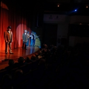 Cena de espetáculo teatral em um palco de madeira iluminado por luz amarela, com cortina vermelha ao fundo. À esquerda, uma atriz usando vestido longo cinza e lenço na cabeça fala ao público. Ao centro, dois atores trajando roupas antigas em tons terrosos estão em posição de fala, com um deles destacado por um foco de luz. Ao fundo, à direita, três músicos vestidos com boinas azuis tocam violão, sanfona e baixo. Na plateia à frente, espectadores assistem atentos em ambiente escuro.