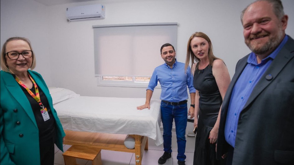 A fotografia mostra quatro pessoas, incluindo o vice-governador Gabriel Souza e a secretária da Saúde, Arita Bergmann, em uma sala de consultório médico. Da esquerda para a direita, Arita Bergmann está sorrindo, usando um blazer verde vibrante. Em seguida, Gabriel Souza, com uma camisa azul, está em pé ao lado de uma maca de exame com lençol branco. Ao seu lado, uma mulher de vestido preto e, por fim, um homem de blazer azul marinho. O ambiente é um espaço clínico novo, com paredes brancas, piso claro e uma janela com persiana.