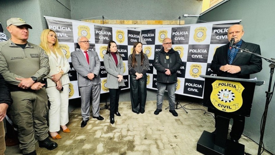 A fotografia mostra um grupo de sete pessoas, incluindo autoridades e representantes, em pé, posando para uma foto de grupo em uma inauguração. Um homem careca, com barba e terno escuro, está no lado direito, falando em um púlpito com a insígnia da Polícia Civil. O grupo atrás dele é composto por um oficial militar com boina, uma mulher de blazer branco, um homem de terno cinza, e duas mulheres com casacos escuros. Eles estão em frente a um banner com o logo e o nome da "Polícia Civil" repetidamente. 