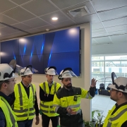 A fotografia mostra um grupo de seis homens, todos usando coletes de segurança amarelos e capacetes de proteção brancos com o logo "CMPC", em um ambiente interno. Eles estão em uma sala moderna, aparentemente uma central de controle, e um deles gesticula enquanto fala com os demais. No fundo, há um grande painel de monitores que exibe uma imagem abstrata em tons de azul. O ambiente é amplo e a iluminação é forte.