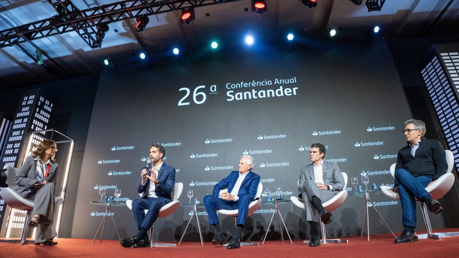 Cinco pessoas participam de um painel em um palco de evento corporativo. Eles estão sentados em cadeiras brancas com fundo escuro onde se lê “26ª Conferência Anual Santander” e vários logotipos do banco. À esquerda, uma mulher com terno cinza segura um tablet e media a conversa. Ao lado dela, quatro homens — todos de terno, exceto o último, que usa suéter e jeans — conversam ao microfone. O ambiente é sofisticado, com iluminação profissional, tapete vermelho e painéis que simulam prédios ao fundo, reforçando o clima urbano e empresarial.