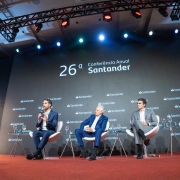 Cinco pessoas participam de um painel em um palco de evento corporativo. Eles estão sentados em cadeiras brancas com fundo escuro onde se lê “26ª Conferência Anual Santander” e vários logotipos do banco. À esquerda, uma mulher com terno cinza segura um tablet e media a conversa. Ao lado dela, quatro homens — todos de terno, exceto o último, que usa suéter e jeans — conversam ao microfone. O ambiente é sofisticado, com iluminação profissional, tapete vermelho e painéis que simulam prédios ao fundo, reforçando o clima urbano e empresarial.