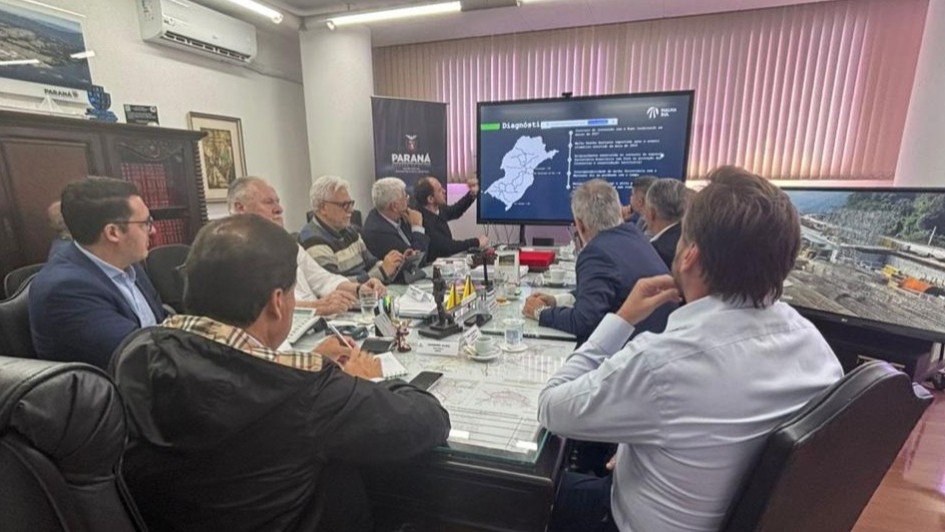 Um grupo de homens está reunido em uma sala de reuniões, sentados em torno de uma grande mesa retangular. Eles observam uma apresentação exibida em uma tela, onde aparece o mapa do estado do Paraná com trechos destacados e informações técnicas. No ambiente, há pastas, papéis, notebooks e microfones sobre a mesa. Atrás do apresentador, há uma cortina fechada e um banner com o logotipo do Governo do Paraná. À direita, uma segunda tela mostra a imagem de uma obra de infraestrutura. A cena transmite uma reunião técnica ou institucional, possivelmente voltada a projetos de transporte ou ferrovias.
