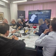 Um grupo de homens está reunido em uma sala de reuniões, sentados em torno de uma grande mesa retangular. Eles observam uma apresentação exibida em uma tela, onde aparece o mapa do estado do Paraná com trechos destacados e informações técnicas. No ambiente, há pastas, papéis, notebooks e microfones sobre a mesa. Atrás do apresentador, há uma cortina fechada e um banner com o logotipo do Governo do Paraná. À direita, uma segunda tela mostra a imagem de uma obra de infraestrutura. A cena transmite uma reunião técnica ou institucional, possivelmente voltada a projetos de transporte ou ferrovias.