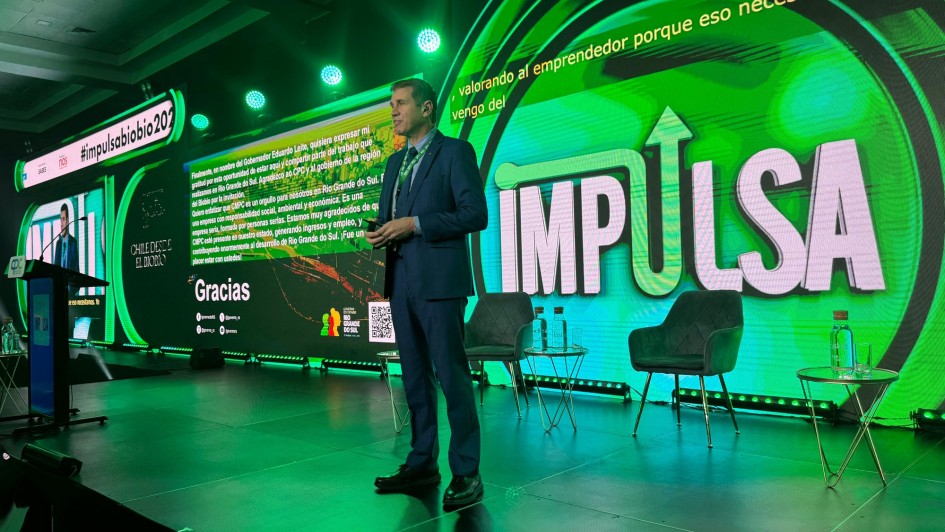 A imagem mostra um palco de evento com iluminação em tons de verde e um telão de LED ao fundo. No centro, um homem branco, vestido com um terno azul-escuro e gravata, está em pé, segurando um microfone. Ele aparenta estar discursando ou fazendo uma apresentação.

Atrás dele, o telão de LED exibe o título "IMPULSA" em letras grandes e brancas, com a palavra "bio" em verde. Ao lado direito, na mesma tela, há a frase "valorando al emprendedor porque eso necesitamos. Yo vengo del" (valorizando o empreendedor porque é disso que precisamos. Eu venho do).

Na parte esquerda do telão, há um texto em português que menciona o Governador Eduardo Leite. O texto diz: "Estamos, em nome do Governador Eduardo Leite, quero expressar mi gratidão por esta oportunidad de estar aquí e agradecer pelo trabalho que tem sido feito no Estado do Rio Grande do Sul e pela região [...] É um privilégio para nós fazermos parte do Rio Grande do Sul. E que estamos orgulhosos de viver em um estado. Uma terra [...] grande, com as pessoas que nós temos. Somos muito agradecidos de fazer parte, gerando riqueza e gerando desenvolvimento. Agradecemos ao desenvolvimento do Rio Grande do Sul." Abaixo deste texto, há a palavra "Gracias" (Obrigado) em letras grandes e brancas.

No canto superior esquerdo, há a hashtag #impulsaBio2025. Ao lado, o nome "CHILE CORTE DE LOS ANDES" (CHILE CORTE DOS ANDES) está escrito em branco.

No palco, à frente do telão, há duas cadeiras de design moderno e duas pequenas mesas de apoio, cada uma com uma garrafa de água e um copo. O chão do palco é preto e brilhante. O ambiente geral é de um evento corporativo ou de inovação.
