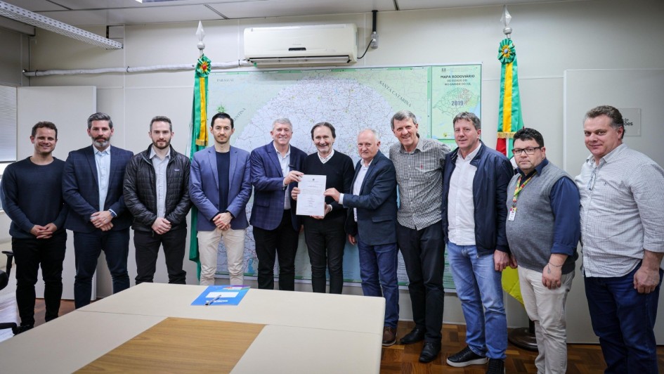 A fotografia mostra um grupo de dez homens em um escritório. O foco está nos dois homens no centro, que estão de terno e seguram um documento em conjunto. Um deles, com uma barba e terno escuro, aperta a mão do outro homem. O grupo de homens ao redor, que vestem desde ternos até roupas casuais com jeans, está olhando para o documento e sorrindo. O ambiente possui paredes brancas e uma grande mesa de reunião em primeiro plano. No fundo, há um mapa rodoviário do Rio Grande do Sul e duas bandeiras.