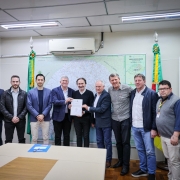 A fotografia mostra um grupo de dez homens em um escritório. O foco está nos dois homens no centro, que estão de terno e seguram um documento em conjunto. Um deles, com uma barba e terno escuro, aperta a mão do outro homem. O grupo de homens ao redor, que vestem desde ternos até roupas casuais com jeans, está olhando para o documento e sorrindo. O ambiente possui paredes brancas e uma grande mesa de reunião em primeiro plano. No fundo, há um mapa rodoviário do Rio Grande do Sul e duas bandeiras.