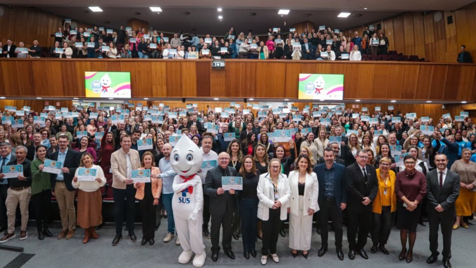 Auditório amplo e lotado, com duas galerias, repleto de pessoas em pé segurando certificados azuis voltados para a câmera. Na frente, um grupo de autoridades posa para a foto, ao lado do mascote Zé Gotinha, símbolo das campanhas de vacinação do SUS. As paredes de madeira e o teto com iluminação clara compõem o ambiente. Ao fundo, dois telões exibem a imagem do Zé Gotinha sobre fundo colorido, reforçando a temática de saúde e vacinação.