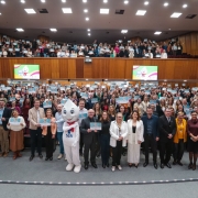 Auditório amplo e lotado, com duas galerias, repleto de pessoas em pé segurando certificados azuis voltados para a câmera. Na frente, um grupo de autoridades posa para a foto, ao lado do mascote Zé Gotinha, símbolo das campanhas de vacinação do SUS. As paredes de madeira e o teto com iluminação clara compõem o ambiente. Ao fundo, dois telões exibem a imagem do Zé Gotinha sobre fundo colorido, reforçando a temática de saúde e vacinação.