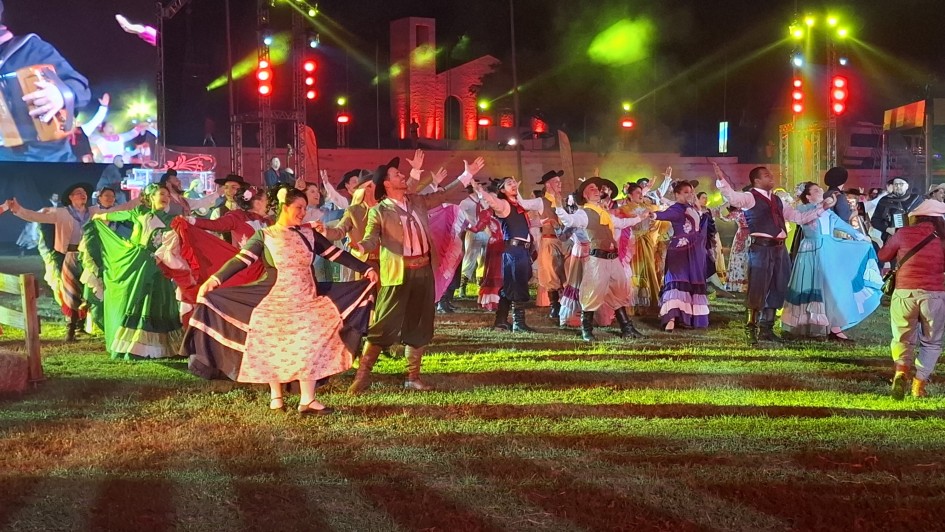 Foto noturna em ambiente aberto, de gramado. Um grupo de mais de 30 pessoas se apresenta com trajes tipicamente gaúchos, com movimentos de dança. O ambiente tem iluminação nas cores verde e vermelho.