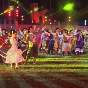Foto noturna em ambiente aberto, de gramado. Um grupo de mais de 30 pessoas se apresenta com trajes tipicamente gaúchos, com movimentos de dança. O ambiente tem iluminação nas cores verde e vermelho.