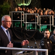 A imagem retrata uma formatura em um auditório, com várias pessoas sentadas em plataformas elevadas ao fundo, decoradas com iluminação verde e elementos suspensos. Em primeiro plano, um homem de terno fala ao microfone em um púlpito, enquanto à direita da imagem estão pessoas em trajes formais e uniformes policiais, sentadas à mesa.