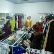 A fotografia retrata o interior de um bazar social. O espaço está repleto de prateleiras e araras com roupas, sapatos e outros itens, organizados para venda. Duas mulheres estão em pé, olhando as roupas em uma arara no centro. Uma delas, de cabelo curto, segura uma blusa verde-limão. Ao fundo, manequins exibem algumas roupas e um ventilador de teto é visível no teto.